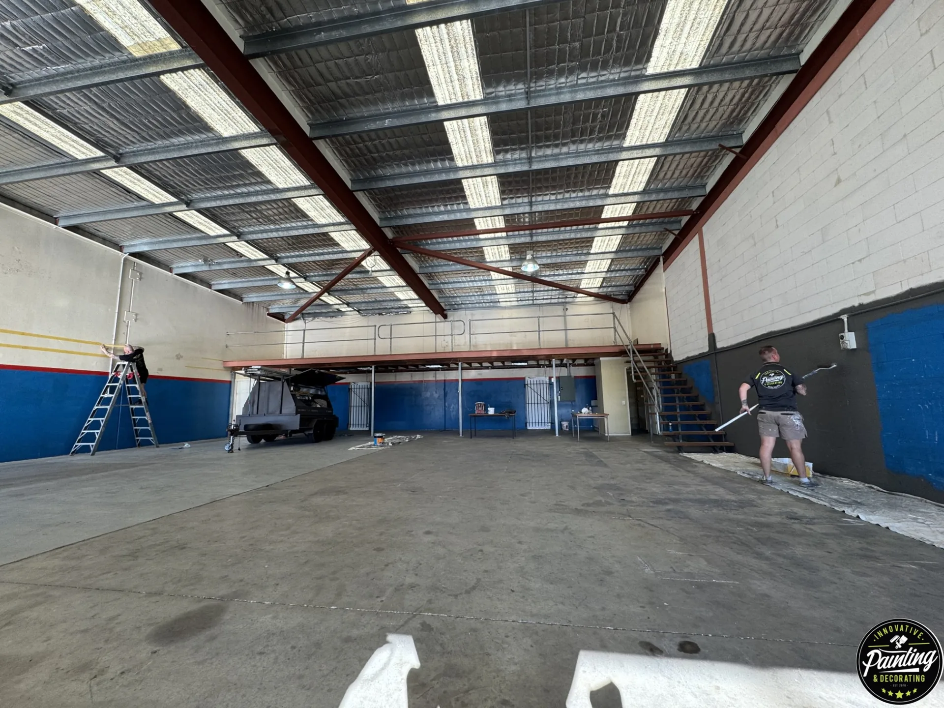 Two painters working on opposite walls inside a large commercial workshop